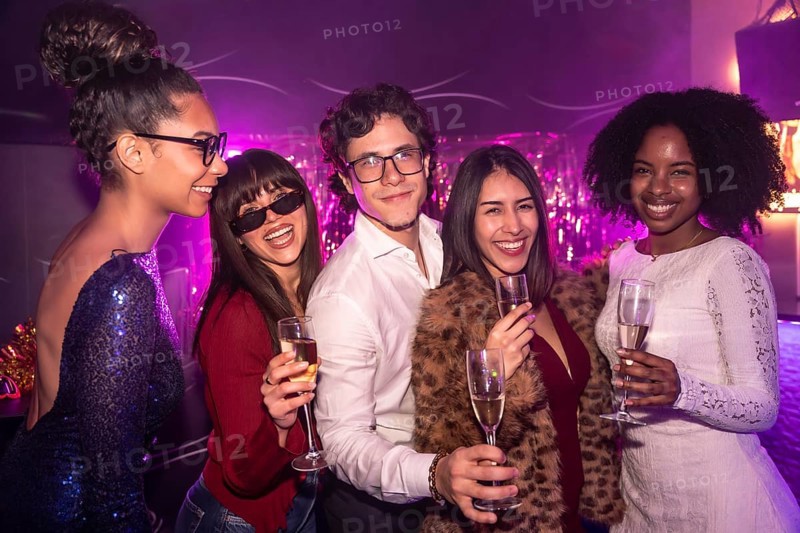 Diverse group of smiling friends celebrating together during a festive event, holding champagne glasses in a lively nightclub with vibrant purple lighting creating a party atmosphere