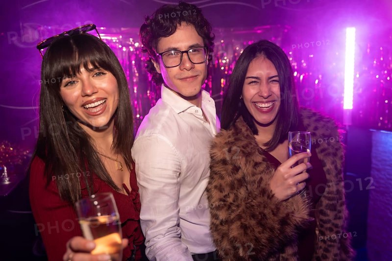 Young friends enjoying a vibrant party atmosphere, smiling and laughing while holding glasses of drinks in a club with purple lights and shiny decorations, embodying joy and nightlife