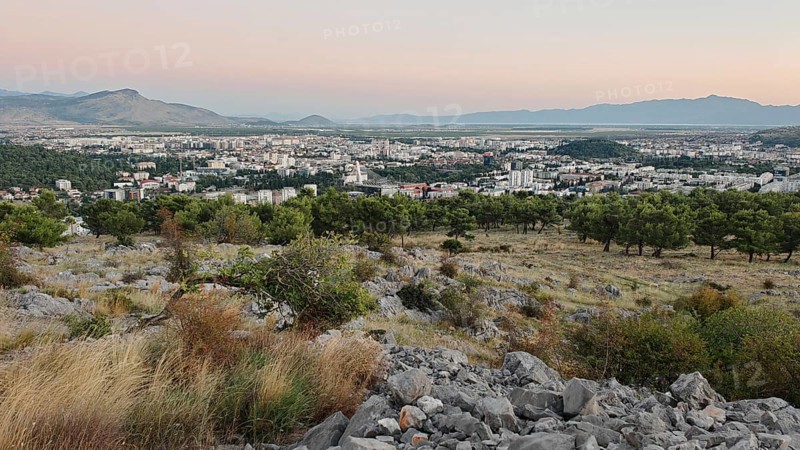 A city surrounded by nature with trees in the foreground and mountains in the background, illuminated by a warm sunset, looking out from a hill on the outskirts of Podgorica, Montenegro