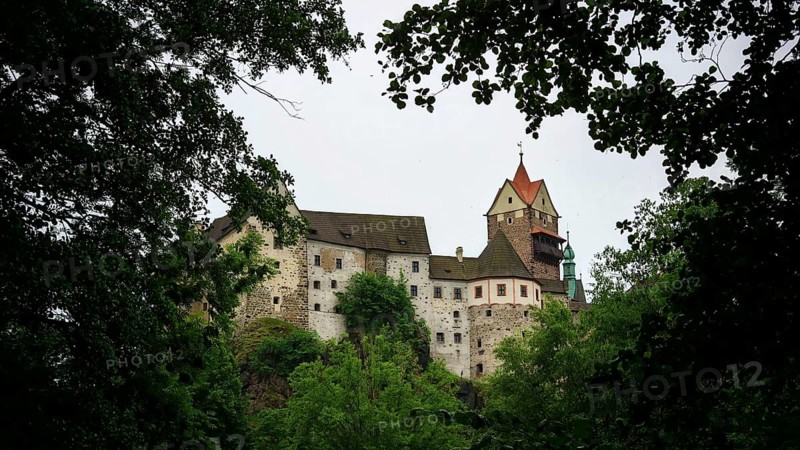 A historic castle rises behind thick greenery under a cloudy sky, Loket, Czech Republic