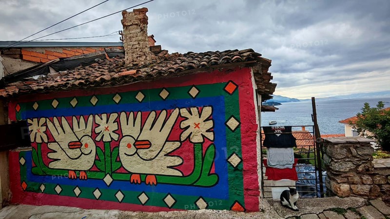 Colourfully painted house façade with dove motifs, laundry hanging to dry with cat in the foreground and lake view in the background, Ohrid, North Macedonia