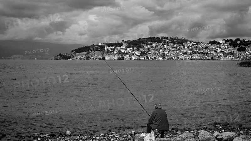Black and white photo of a fisherman on a lake shore with a city in the background, Ohrid, North Macedonia
