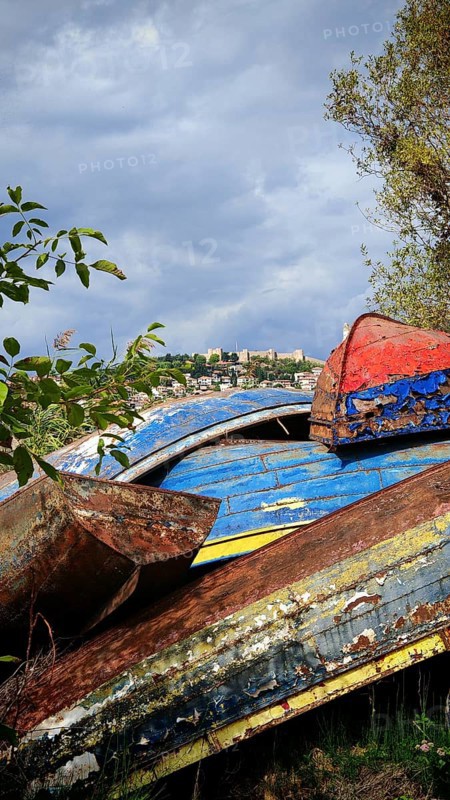 Weathered, colorful boats lie under a cloudy sky surrounded by trees and shrubs, Lake Ohrid, North Macedonia