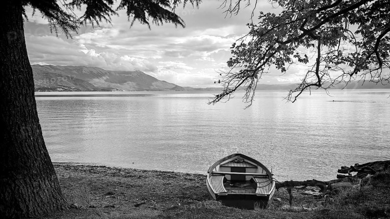 A boat on the shore of a calm lake surrounded by trees in black and white, Lake Ohrid, North Macedonia
