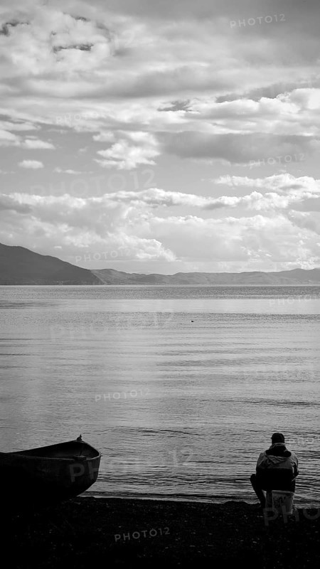 An angler is quietly looking at the shore of a lake, accompanied by a boat in black and white, Lake Ohrid, North Macedonia