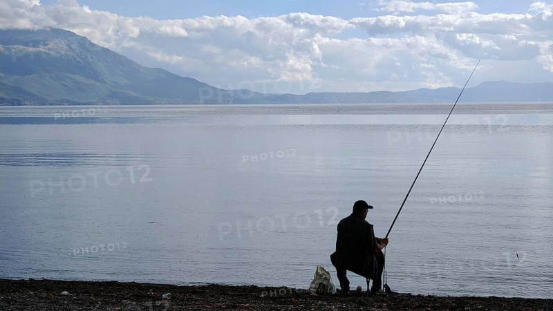 An angler sits on the shore of a peaceful lake, surrounded by mountains and a peaceful, natural atmosphere, Lake Ohrid, North Macedonia