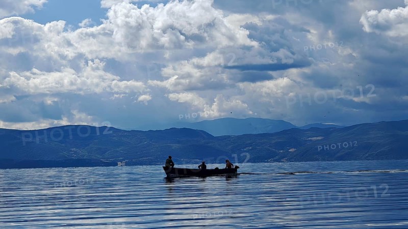 A small boat with silhouettes in a calm lake surrounded by mountains and dramatic cloud formations, Lake Ohrid, North Macedonia