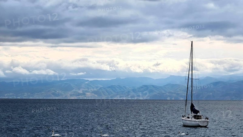 A sailboat on a calm sea with mountains in the background and cloudy sky, Lake Ohrid, North Macedonia