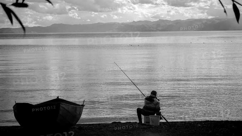An fisherman sitting on the shore of a calm lake, with a small boat and a black and white aesthetic, Lake Ohrid, North Macedonia