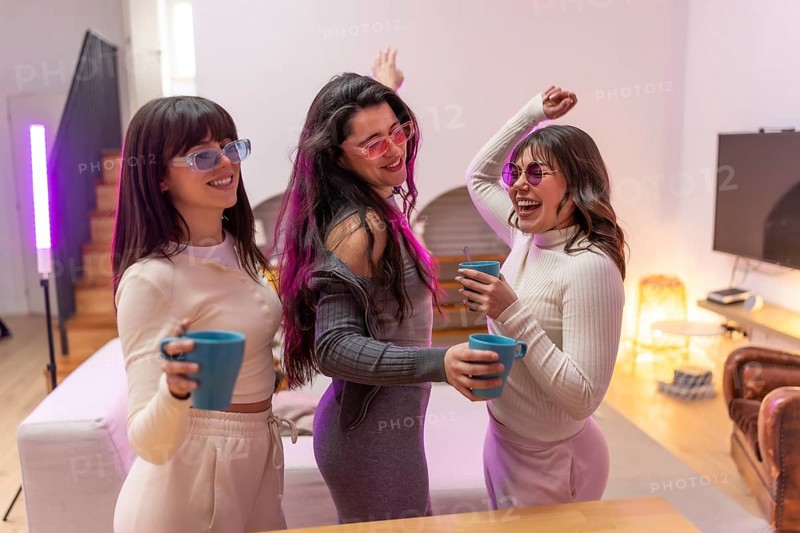 Three happy young women friends wearing sunglasses and casual clothes dancing, holding mugs, and laughing together while enjoying a home party with vibrant neon lights
