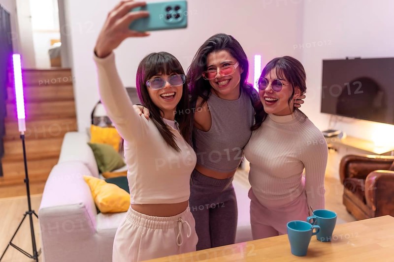 Three cheerful young women wearing colorful sunglasses gathering for a selfie in a modern living room with atmospheric lighting, enjoying a fun moment together