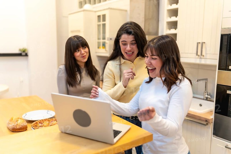 Group of female friends gathering in a modern kitchen, sharing an exciting moment while celebrating a victory, success, or positive news seen on a laptop screen