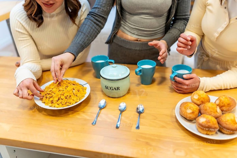 Three young women gather around a wooden table, preparing a morning breakfast with a bowl of cornflakes, freshly baked muffins, a sugar bowl, and cups of milk, enjoying togetherness