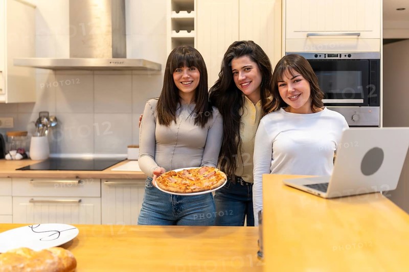 Three young women smiling and looking at camera while one is holding a freshly baked pizza in a modern kitchen, sharing a happy moment together at home