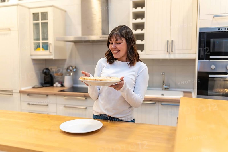 Young woman smiling in a modern kitchen while holding a freshly baked pizza on a plate, enjoying homemade cooking and domestic life, preparing a casual dinner with joy and pride