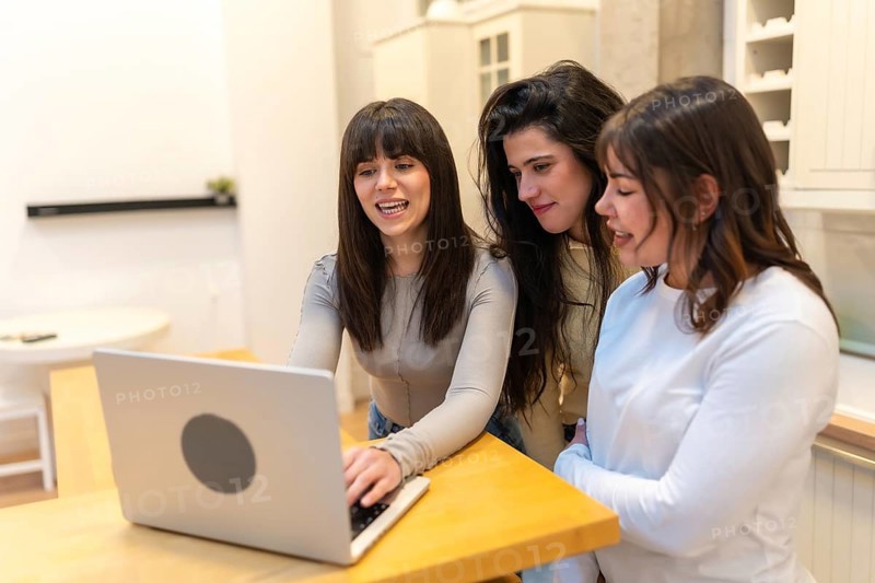 Three friends, women, collaborating and smiling while looking at a laptop computer on a wooden counter, sharing a moment of remote work or online connection in a home setting