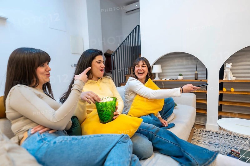 Three women on a sofa sharing popcorn, laughing and watching tv together in a cozy living room, enjoying a relaxed movie night and casual friendship at home