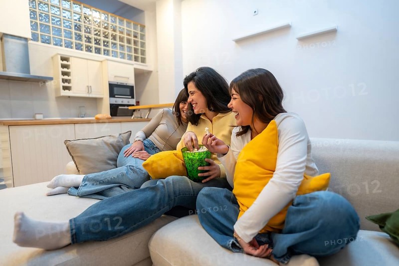 Group of diverse young women enjoying a relaxed evening on the couch at home, sharing laughter and popcorn while watching television together creating a bonding moment