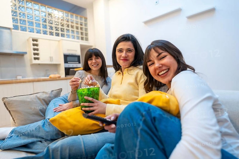 Three young women laughing and relaxed on a couch at home, enjoying a cozy movie night together with popcorn, streaming a film and bonding in casual clothing