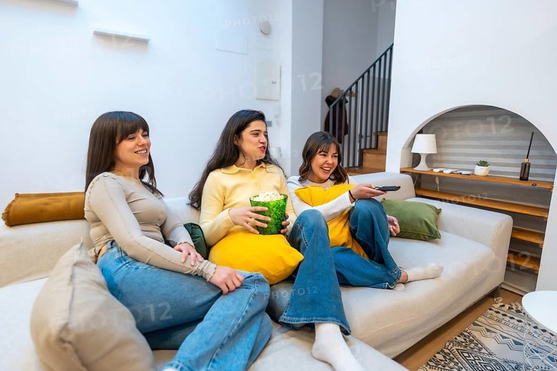 Group of happy young women friends relaxing on a comfortable couch at home, spending leisure time together, laughing and watching tv during a cozy movie night with popcorn