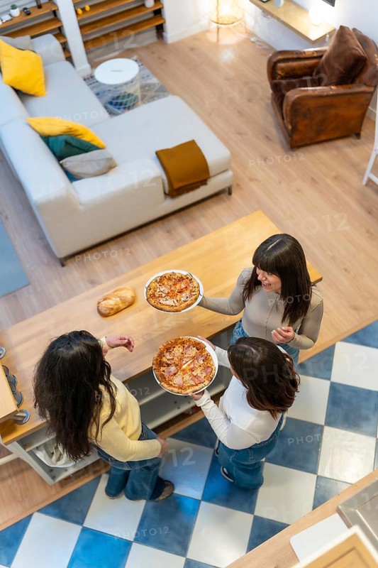 Three happy young women gathered around a wooden kitchen island sharing homemade pizza, chatting and laughing during a relaxed, casual get together at home