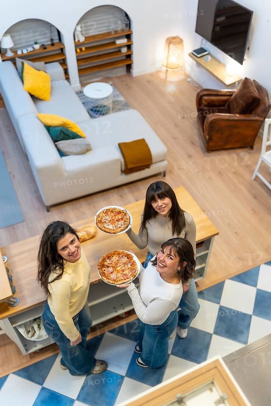 Three cheerful young women standing in an open plan apartment, holding pizzas and looking up while smiling, showing friendship and enjoying domestic leisure
