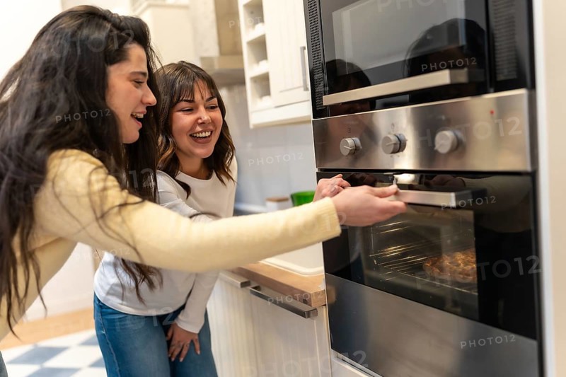 Two cheerful women are laughing together while one opens the oven door to check on a pizza baking inside, creating a warm atmosphere of friendship and domestic happiness