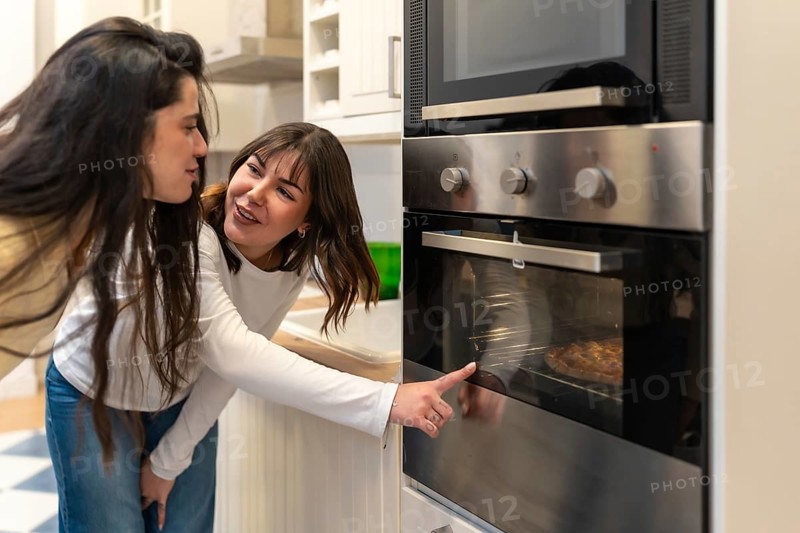 Two women in a modern kitchen lean over an electric oven, checking a homemade pizza together, sharing smiles and casual conversation in a warm, domestic moment
