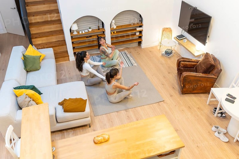 Three young women smiling and doing yoga on a mat in their modern living room, prioritizing wellness, fitness, and friendship during a home workout session