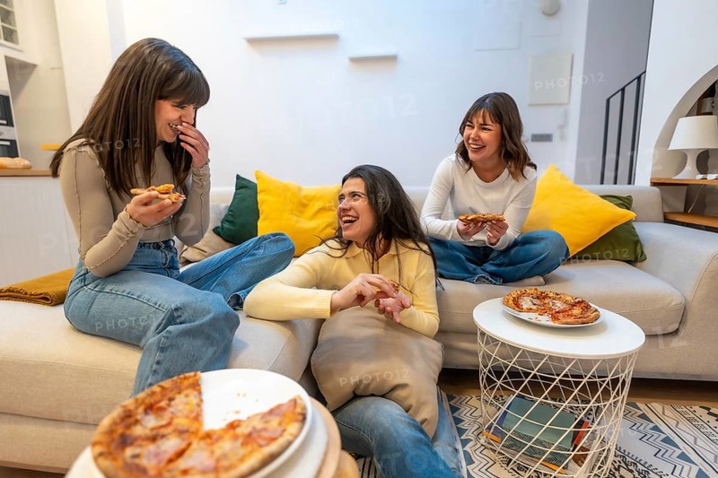 Three young women relaxing on a couch and floor, sharing pizza slices during a casual gathering, enjoying friendship and candid moments of laughter indoors