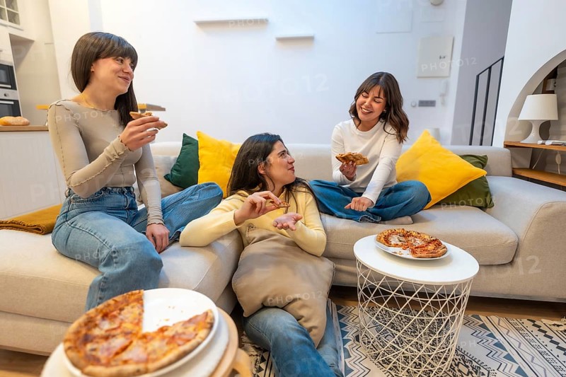 Three young women laughing and talking while eating pizza slices on a sofa in a modern living room, sharing a leisurely meal and friendship during a relaxed gathering
