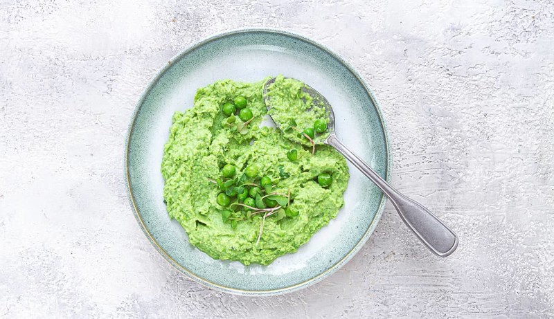 A vibrant green pea puree fills a shallow bowl, topped with fresh peas and microgreens. The dish looks fresh and inviting, ideal for a light, nutritious meal at any time