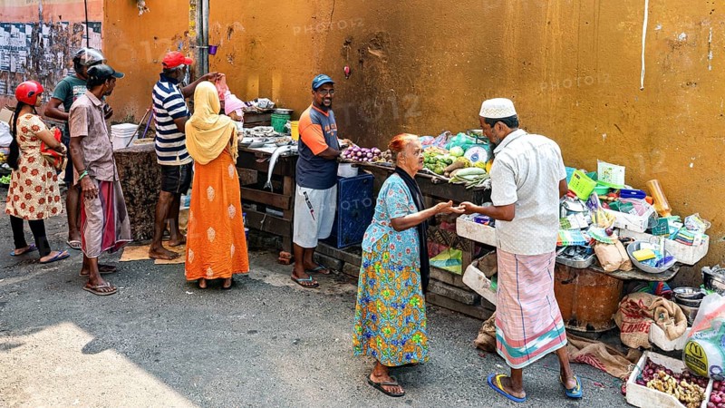 People trade and interact at a market stand full of fresh vegetables and goods