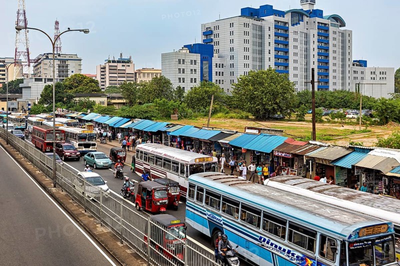 City street with several buses and market stalls, surrounded by large buildings and trees, buses in traffic from Colombo in Sri Lanka