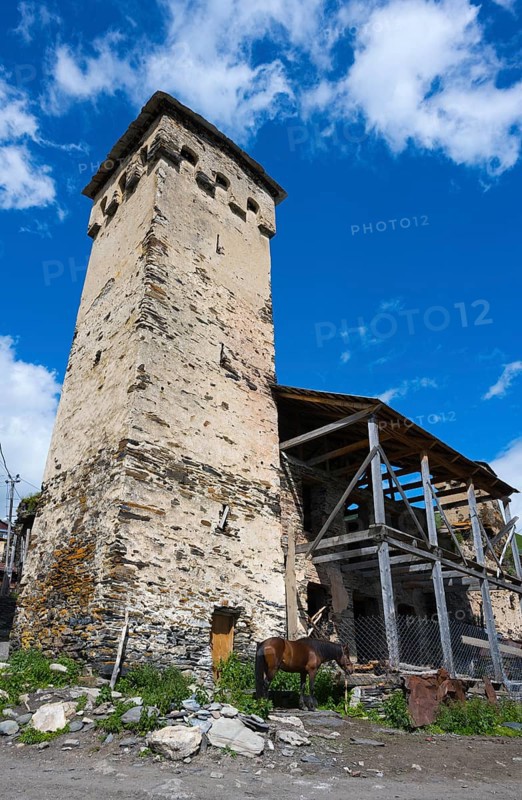 Tall stone building next to a wooden shelter with a horse against a blue sky. Rural landscape, defensive defence tower, Ushguli, Ushguli, Shibiani district, Zhibiani, Upper Svaneti, Svaneti, Georgia