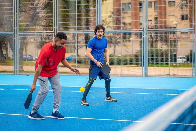 Two diverse young men compete on a blue outdoor pickleball court, focused and energetic as they rally with paddles under sunny urban skies, enjoying sport and fitness together
