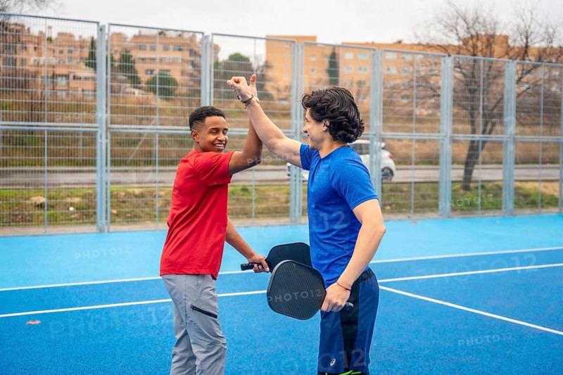 Two smiling male athletes, one african american and one caucasian, giving a high five on a blue outdoor pickleball court, celebrating teamwork and good sportsmanship after a game