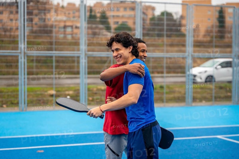 Two diverse male friends smiling and hugging on a blue pickleball court after a game, demonstrating companionship, teamwork, and healthy competition in an urban setting