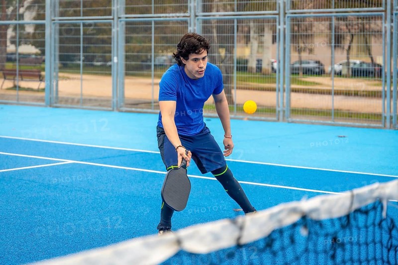 Young man concentrating on a pickleball match on a blue outdoor court, holding his paddle ready to hit the yellow ball over the net, focused and athletic in action
