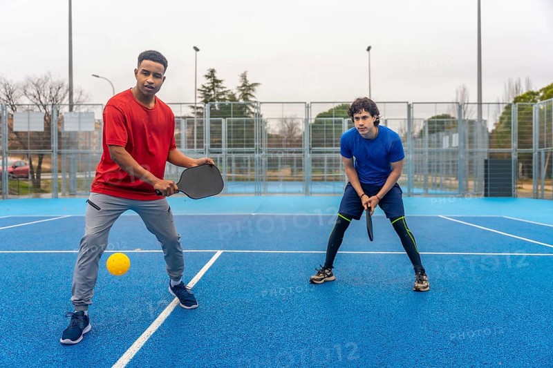 Two young men actively engaged in a pickleball game on an outdoor blue court, holding rackets and preparing to hit the ball during competitive sports activity