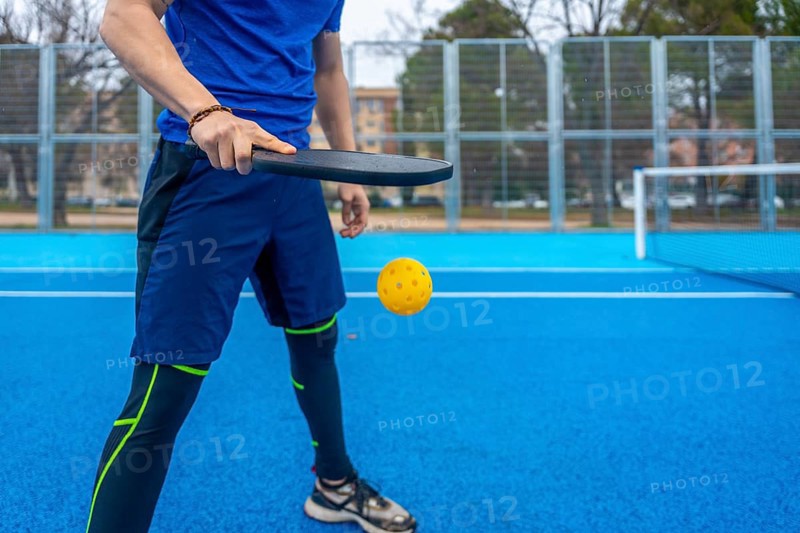 Sportsman wearing athletic apparel, playing pickleball with a paddle and yellow plastic ball on an outdoor vibrant blue court during a competitive match or practice session