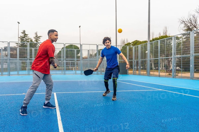 Two young menone african american, one caucasiancompete in an energetic outdoor pickleball rally, paddles in hand on a bright blue court, showcasing fitness and friendship