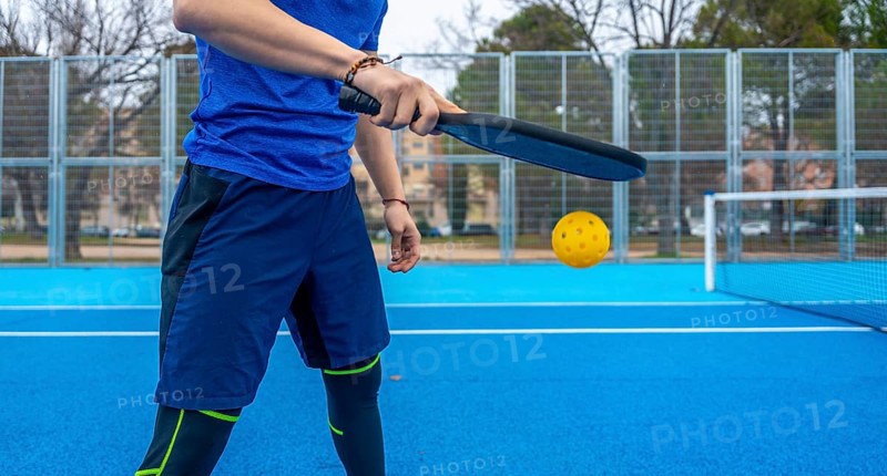 Athlete striking a yellow pickleball with a paddle on a vibrant blue outdoor court, mid rally showcasing focus, power and summer fitness during casual competition