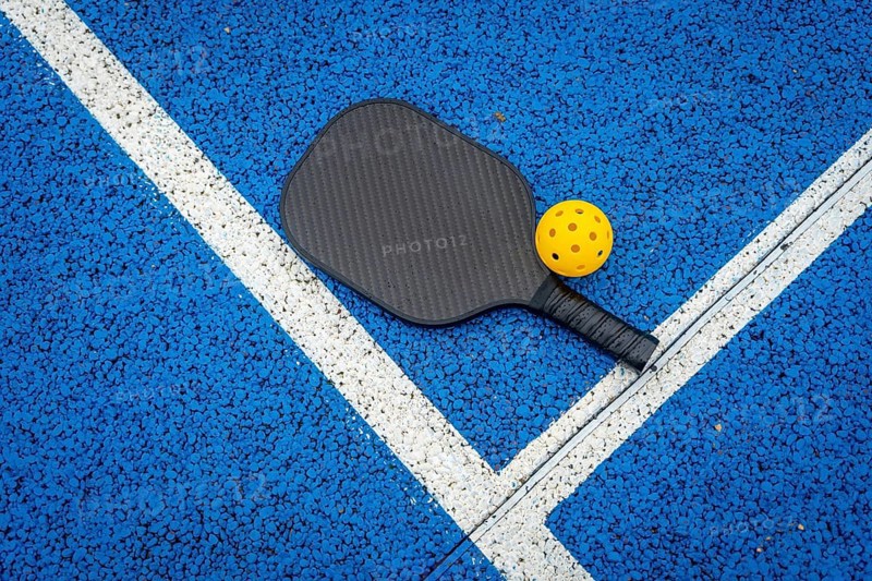 Pickleball paddle and yellow wiffle ball resting on the vibrant blue court surface with distinct white boundary lines, representing an active lifestyle and recreational sport