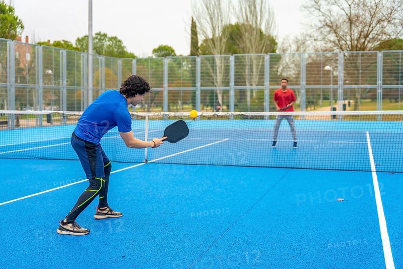 Two young men actively engaging in a game of pickleball, one hitting the yellow ball with a paddle, on a bright blue outdoor court with a net, enjoying leisure and fitness