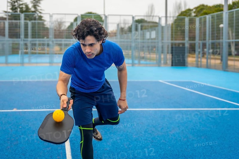 Male athlete concentrating while holding a pickleball paddle and yellow ball, ready to serve or return on a vibrant blue outdoor court, engaging in active sport and fitness