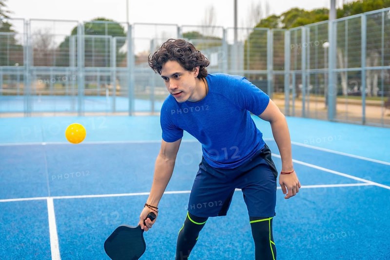 Young man holding a pickleball paddle, focused and ready to hit the yellow ball during an outdoor sports game on a vibrant blue court, representing fitness and recreation