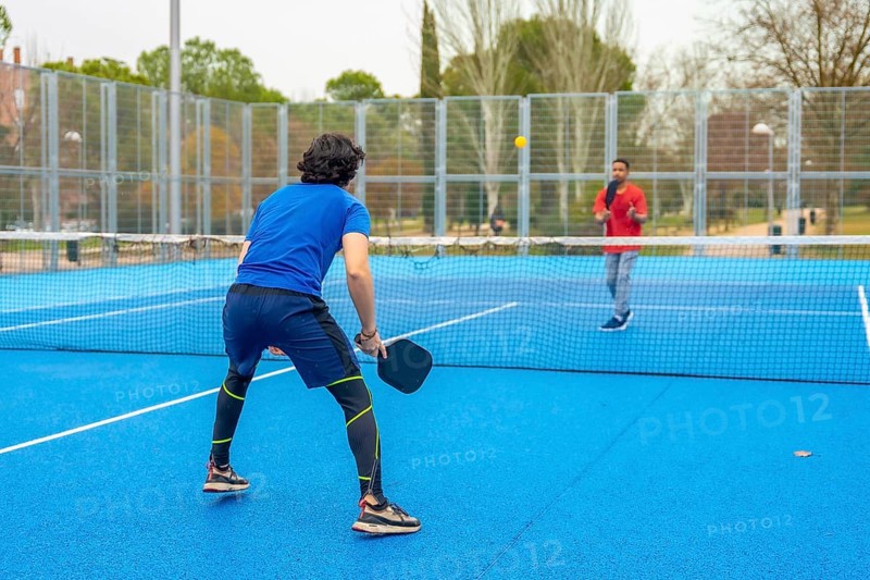 Two male athletes playing an intense pickleball game on a vibrant blue court, holding paddles and focusing on hitting the ball over the net during a competitive and active sporting event outdoors
