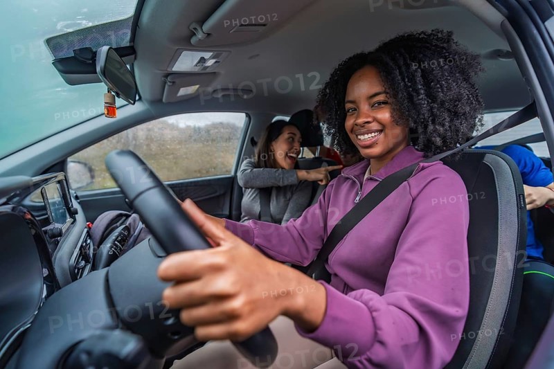 Smiling young black woman driving a car with friends in the back, laughing and pointing, capturing the joyful essence of a group road trip and travel for sports