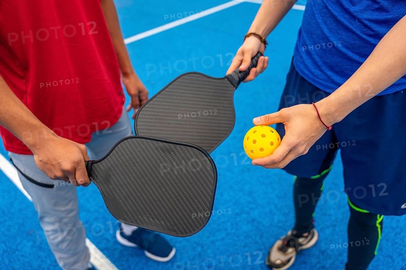 Two people are ready to play pickleball, holding paddles and a bright yellow wiffle ball on a vibrant blue court, highlighting recreation and active lifestyle